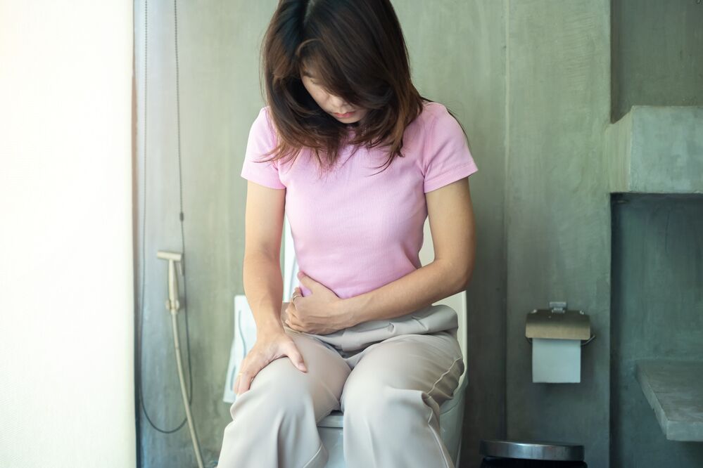 Young woman fully clothed sitting on a toilet with the lid down holding her stomach to help illustrate How Long Before You Poop After Gastric Sleeve